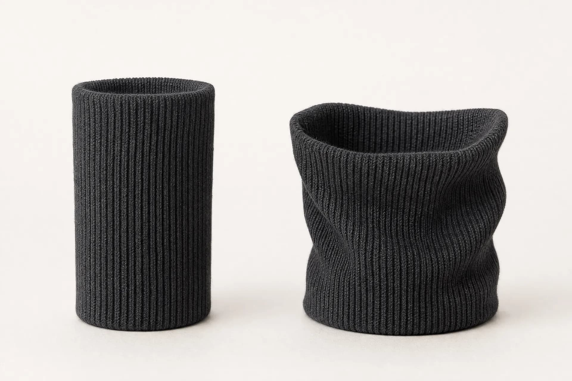 Two identical graphite ribbed sock cuffs standing side by side: the left holds a narrow even cylinder, the right has slumped wider and slacker at the mouth.